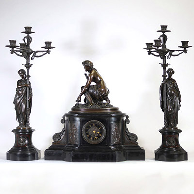 Garniture de cheminée en bronze patiné et marbre noir, signée Delvaulx, époque Napoléon III Imposante garniture de cheminée en bronze patiné et marbre noir composée d’une pendule centrale surmontée d’une figure féminine nue, assise dans une posture de toilette, finement sculptée et reposant sur un socle tripartite orné de bas-reliefs à sujet antique. Elle est accompagnée d’une paire de candélabres